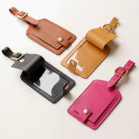 Four leather luggage tags in brown, black, beige, and pink on a light gray background.