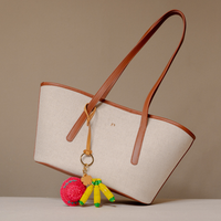 A structured tote bag made of light beige canvas with brown leather trim and handles, featuring a small gold logo on the front. A decorative keychain with a pink pom-pom and yellow tassels hangs from the bag.