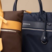 Two crossbody bags, messenger bags, one brown and yellow, ballistic nylon with leather details and handle, one brown with yellow and the other navy with 'DANILOS' branding, on a beige background.