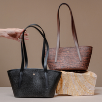Two woven tote bags with long handles are shown on a beige studio background. A hand holds the black bag in the foreground, while a second brown woven bag sits behind it on a stone block.