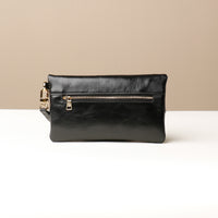 Front view of a glossy black leather Karelia Clutch Danilos with a gold zipper over a white and beige background